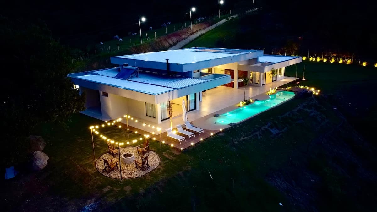 Vista aérea noturna da casa iluminada com piscina e fogo de chão